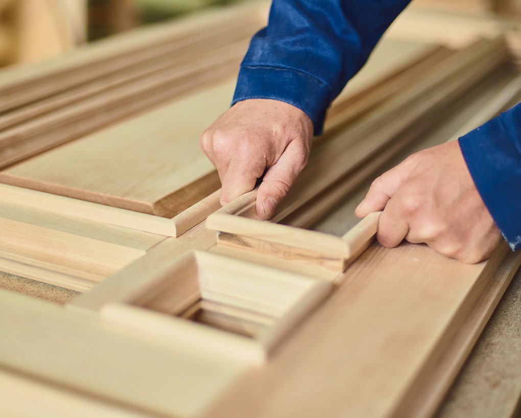 Due mani che lavorano il legno, posizionando e assemblando pezzi di pregiato materiale in un ambiente artigianale. Lo sfondo mostra vari pannelli di legno.
